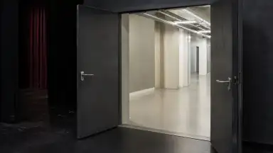En teaterdør står på klem mellem et mørkt scenerum og en klart oplyst gang med synligt håndtag på begge sider.