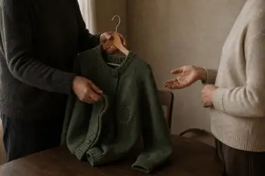 To voksne mødes om en repareret mørkegrøn cardigan på en bøjle; den ene viser reparationen, mens den anden svarer med en åben hånd.