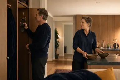 En voksen hænger en aftagelig manchet i et åbent skab, mens en anden stiller to skåle frem i køkkenet.